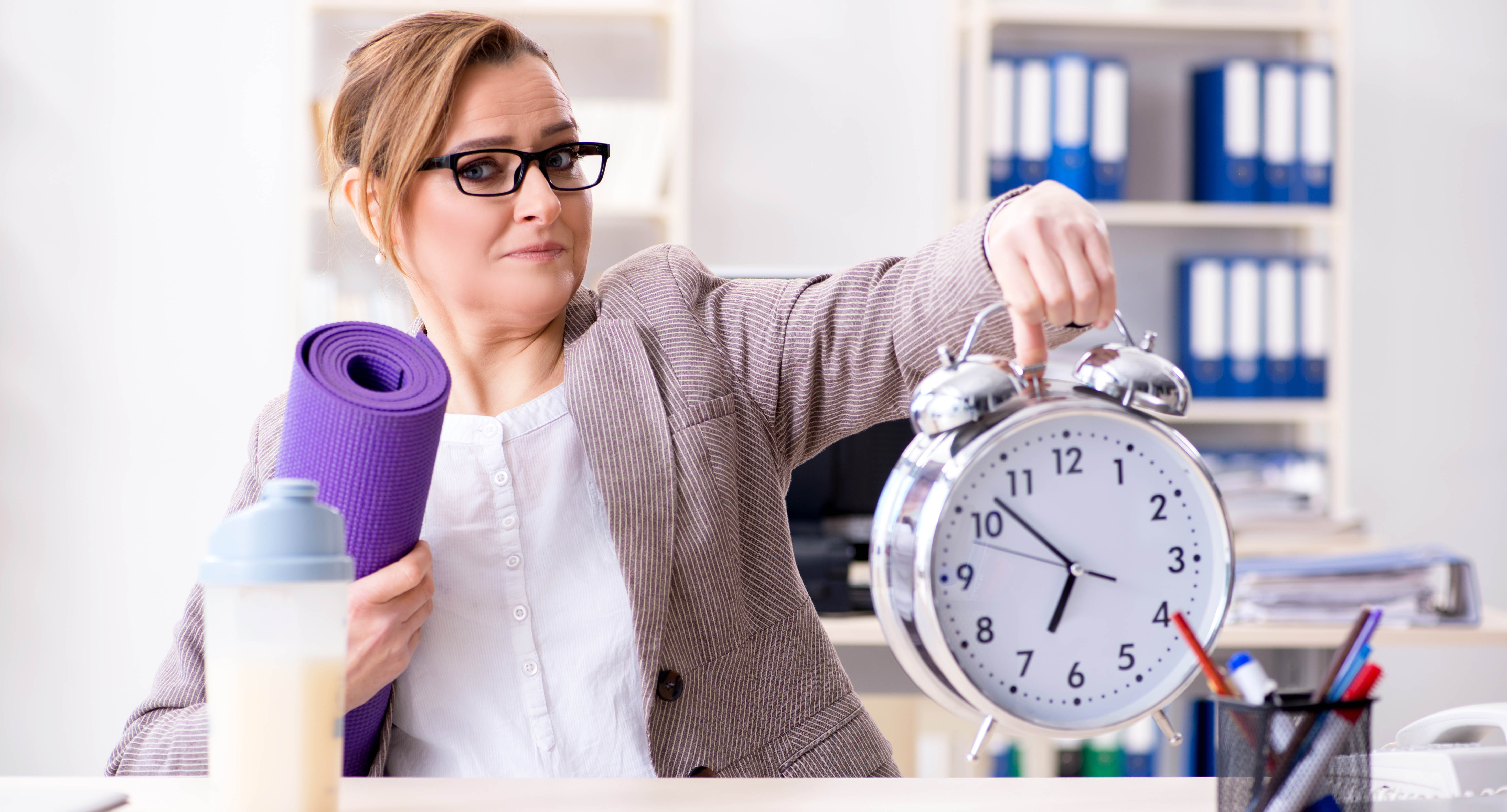 Professional woman sitting at her desk holding a large alarm clock in one hand and her purple yoga mat in the other, preparing to go to the gym for a workout Professional woman sitting at her desk holding a large alarm clock in one hand and her purple yoga mat in the other, preparing to go to the gym for a workout